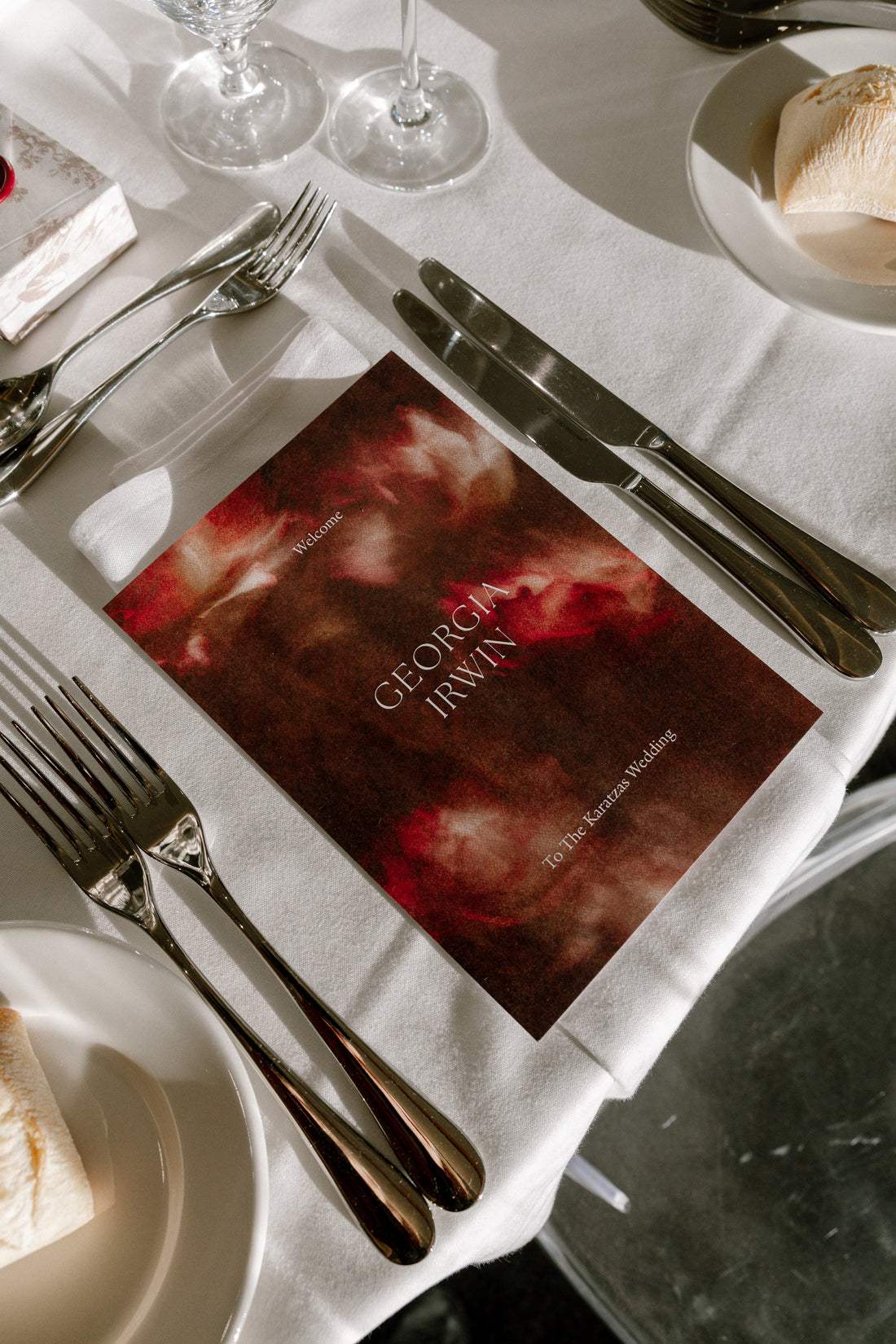 A burgundy floral wedding menu card with white serif lettering, styled on a white linen napkin beside cutlery at a reception table